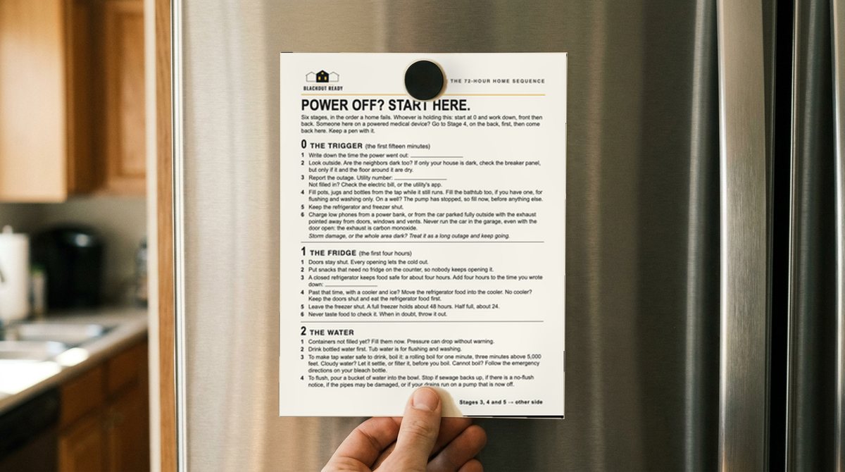 A hand holding the Blackout Ready fridge-door card flat against a refrigerator door under a magnet, in an ordinary kitchen in daylight.
