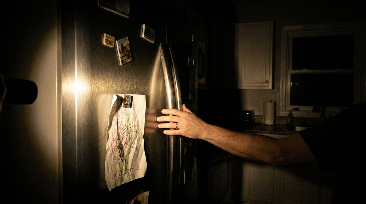 A closed refrigerator door in a dark kitchen at night, lit by a flashlight from out of frame, a hand resting on the handle.