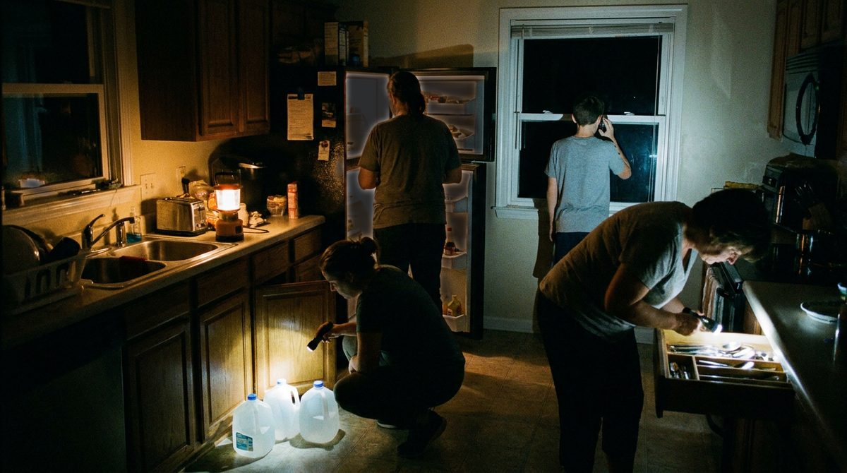 A dark kitchen at night lit by one lantern and flashlights. Four family members are busy at once: one at the open refrigerator, one counting water jugs at a cupboard, one on the phone at the window, one searching a drawer.