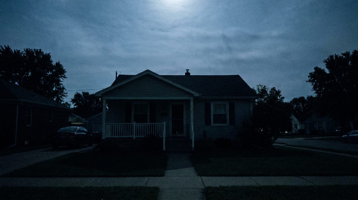 The front of an ordinary house at night during an outage: porch light off, every window dark, the neighboring houses dark, faint moonlight.